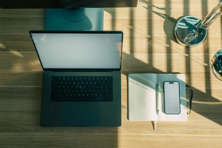 Laptop, phone and notebook on a sunlit wooden desk